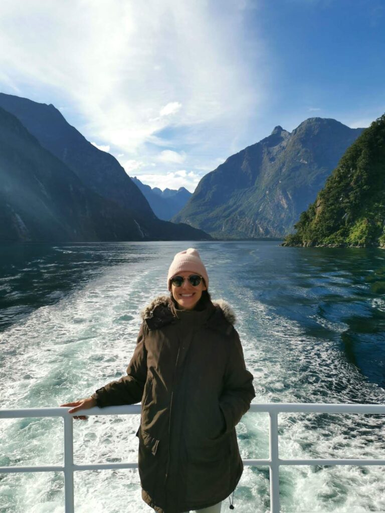 Katharina smiling on the deck of a cruise boat in Milford Sound with deep blue water and towering mountains in the background.
