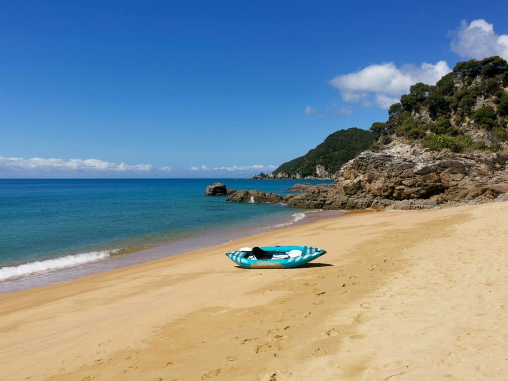 A blue and white inflatable kayak sits on the golden sand of a quiet beach in Abel Tasman National Park, with calm turquoise water and rocky outcrops under a clear blue sky.