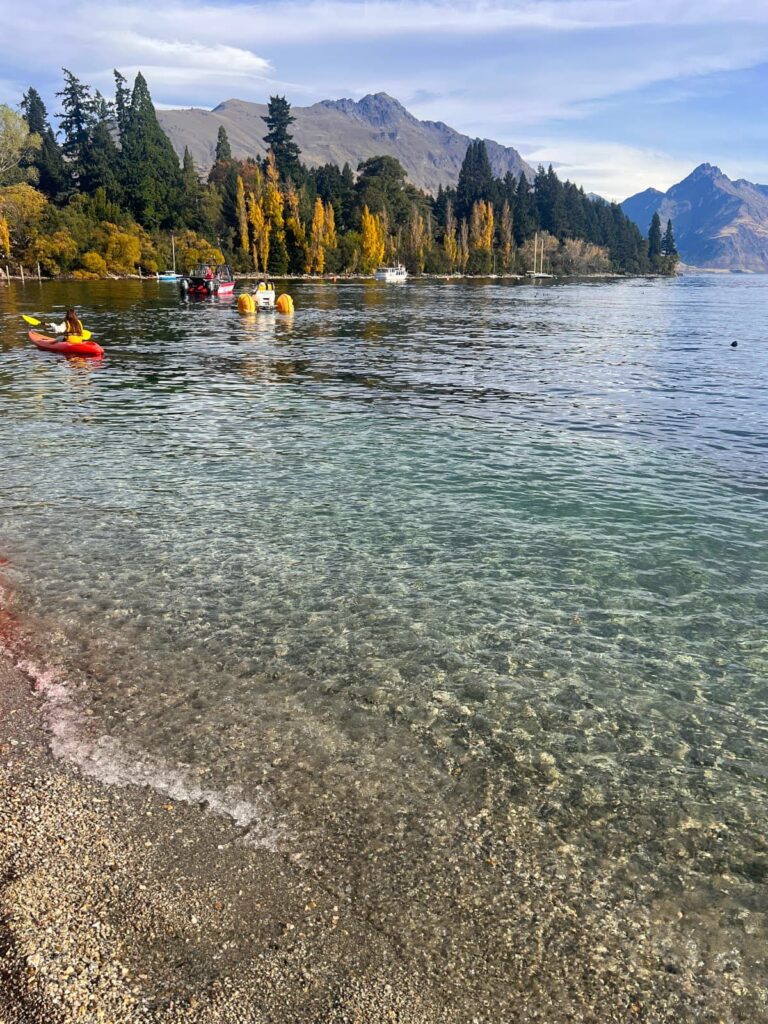 People kayaking on the clear, calm water of Lake Wakatipu in Queenstown with pine trees and mountains in the background.