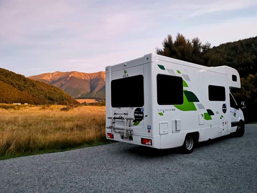 A white KEA campervan parked at Teetotal Campsite, with a golden grassy field and mountains glowing in the distance under a dusky sky, near Lake Rotoiti, New Zealand.