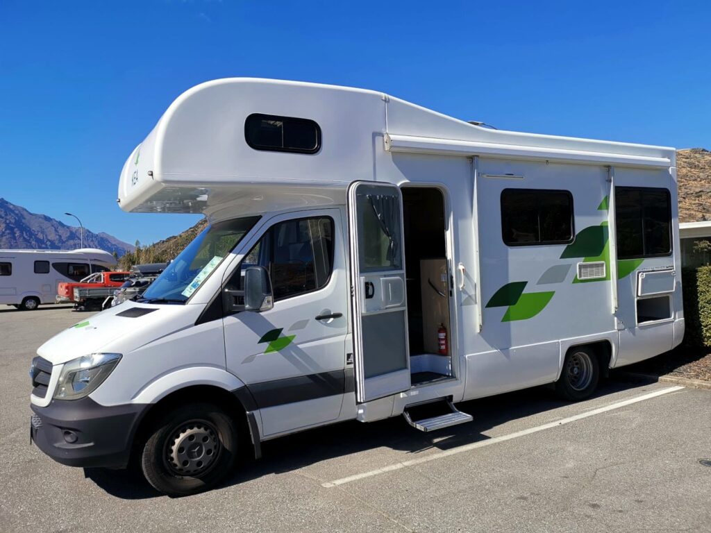 A large white Kea Platinum campervan parked in a sunny paved lot at Queenstown airport with its side door open. A perfect home on wheels for a South Island trip.