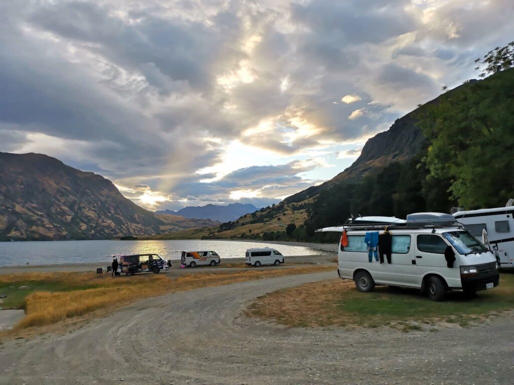 Several campervans parked at the Kidds Bush campsite on the shore of Lake Hāwea, with mountains and a cloudy sky at sunset in the background.