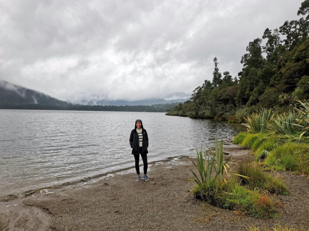 Katharina standing on the gravel shore of Lake Paringa's Department of Conservation campsite under a cloudy sky, surrounded by dense native forest.