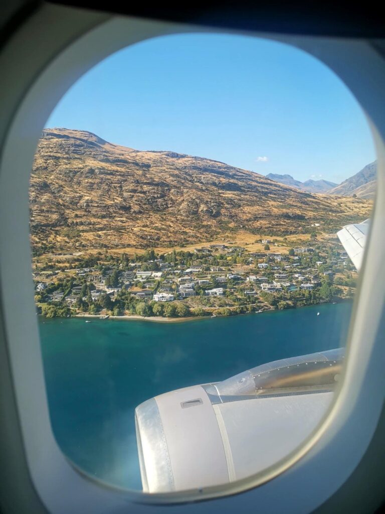 A view from a plane window looking down at houses along the turquoise water of Lake Wakatipu and the golden hills of Queenstown which is a perfect start for a 2-week campervan itinerary.