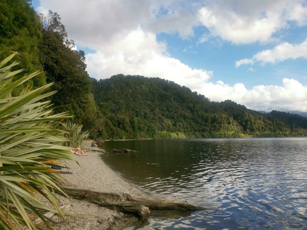 View from a pepple beach at Lake Mapourika's Otto/MacDonalds Campsite, looking across the calm water toward dense green forest and hills under a partly cloudy sky.