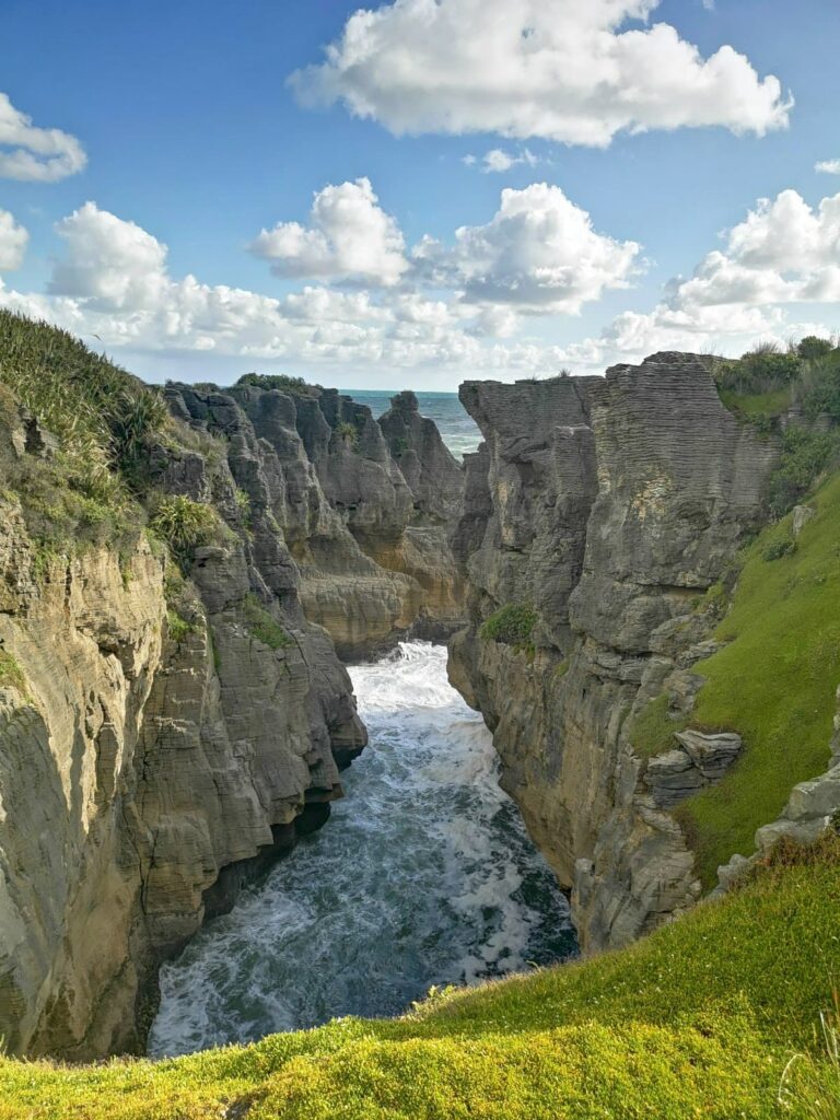 A view looking down into a narrow surge pool between high limestone cliffs at Pancake Rocks, where white waves crash against the rock walls under a bright, cloud-filled sky.