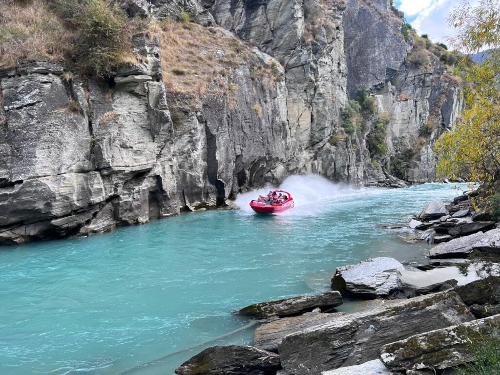 A red jet boat speeding through a narrow rocky canyon on the bright blue Shotover River, one of the best things to do in Queenstown.