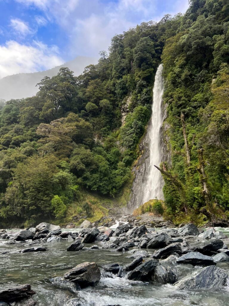A tall, thin waterfall plunging into a rocky river surrounded by a dense green rainforest.