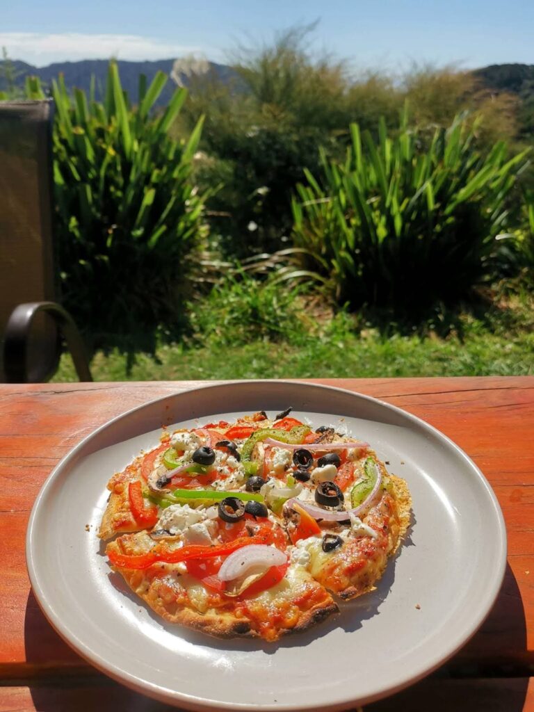 A thin-crust vegetable pizza from Totos Pizzeria topped with olives, peppers, onions, and feta cheese sits on a white plate on a wooden table, with a scenic view of green hills and flax plants in the background.