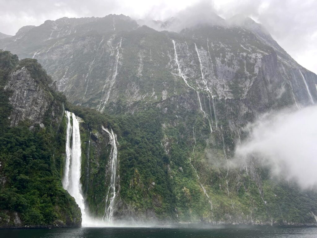 Multiple waterfalls flowing down high, misty green cliffs into the dark water of Milford Sound on a rainy and cloudy day.