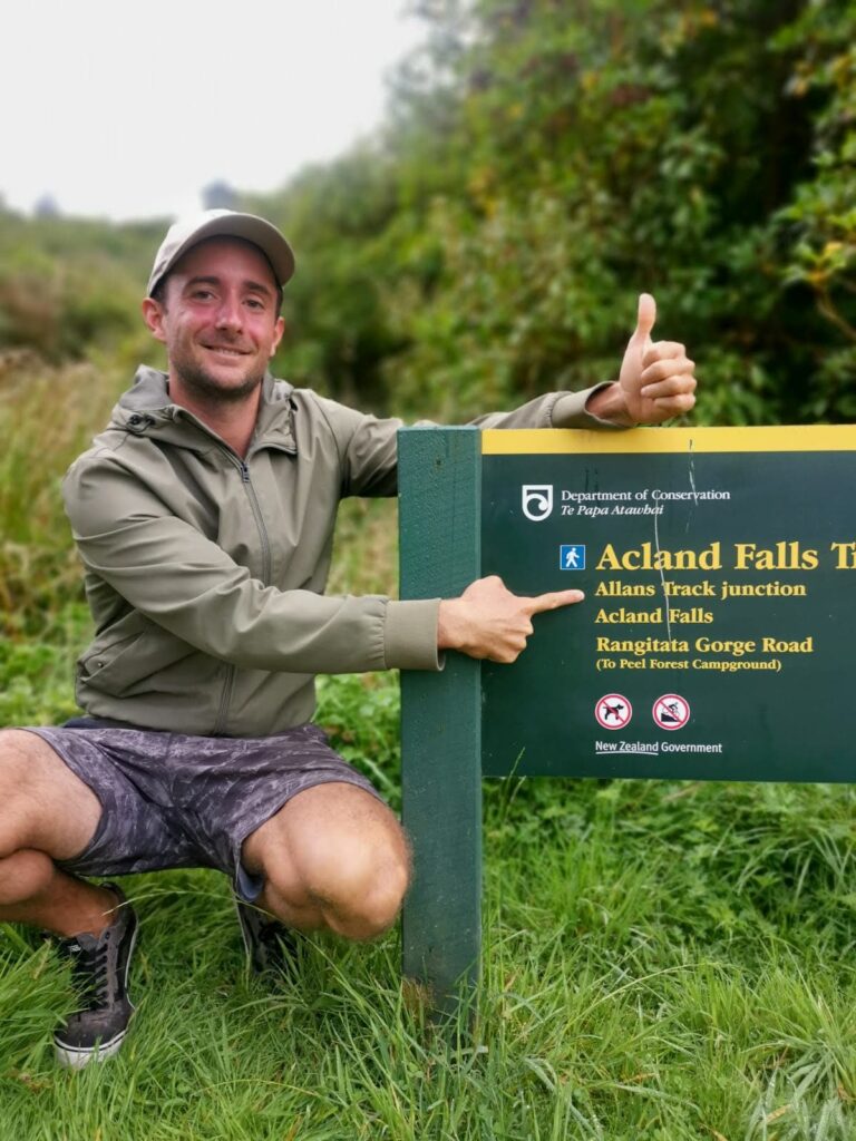 Allan gives a thumbs up while pointing to "Allans Track junction" on a Department of Conservation sign for the Acland Falls Track in Peel Forest.