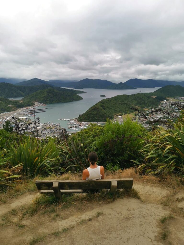 Katharina sitting on a wooden bench overlooking the town of Picton and its harbor, with the rolling green hills and waterways of the Marlborough Sounds extending into the distance under a cloudy sky.