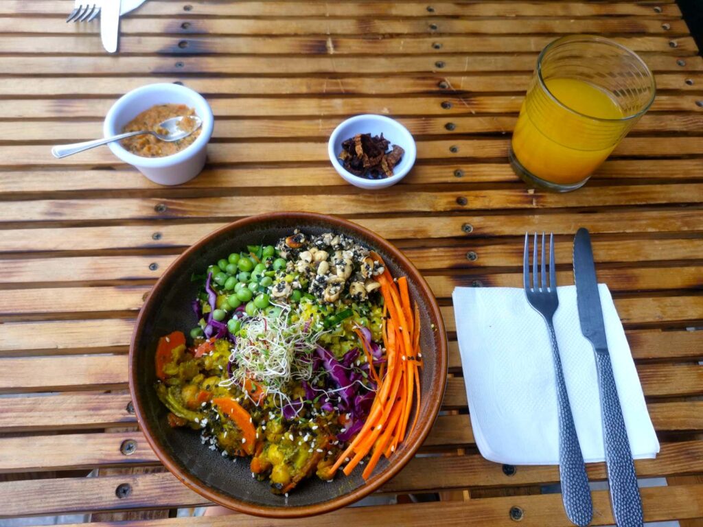 A colorful veggie bowl sitting on top of a wooden table with two small containers of condiments and a glass of orange juice behind it