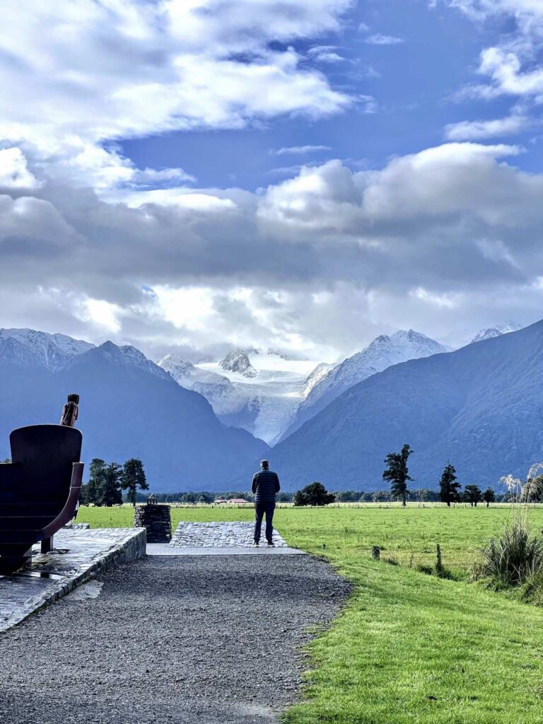 A man stands at the end of a gravel path with a view of fox glacier plunging down the southern alps in the distance with green grassy fields in the foreground