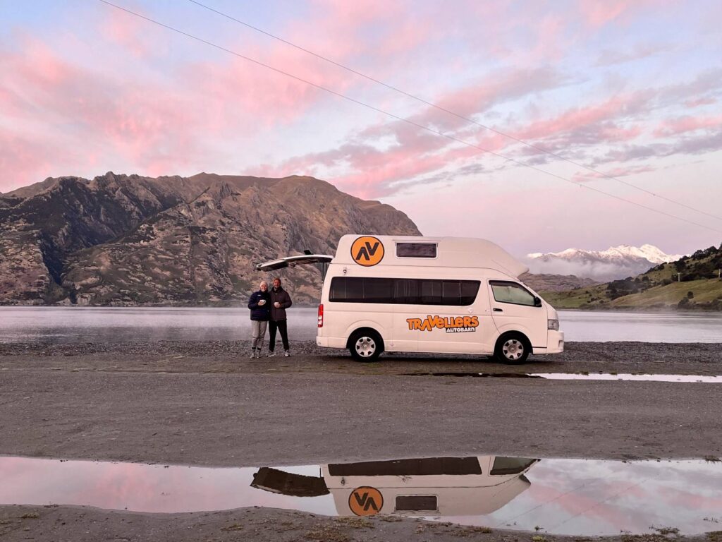 A white Travellers Autobarn campervan parked on the lakefront at Kidds Bush Campsite during a vibrant pink sunrise, a highlight of a South Island New Zealand itinerary. A man and woman stand together holding coffee mugs, with the van and mountains reflected perfectly in a puddle in the foreground.