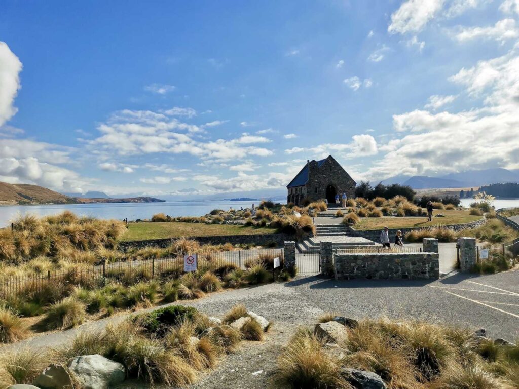 The historic stone Church of the Good Shepherd sitting amongst golden tussock grass on the shore of Lake Tekapo under a partly cloudy blue sky.