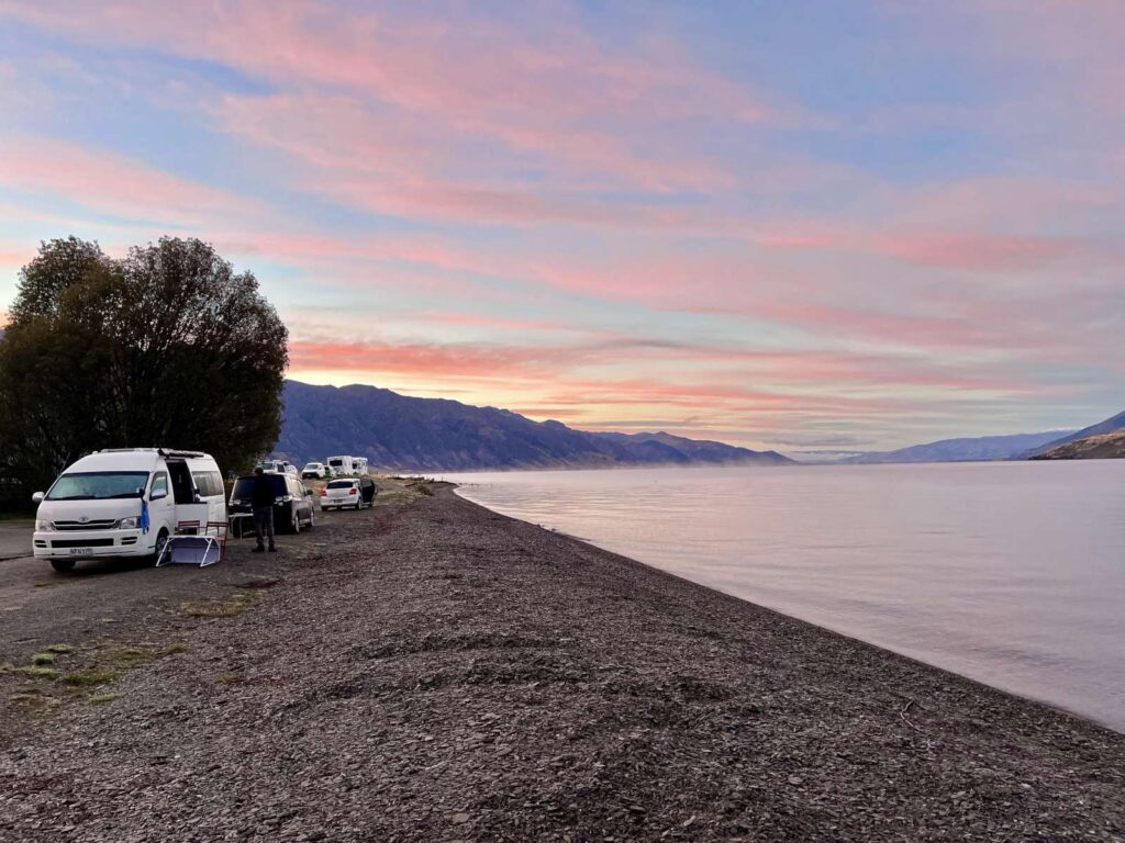 Several campervans and cars parked along the gravel shoreline of Lake Hāwea at Kidds Bush Campsite during a vibrant pink and orange sunrise, a popular free or low-cost camping stop included on a South Island New Zealand itinerary.