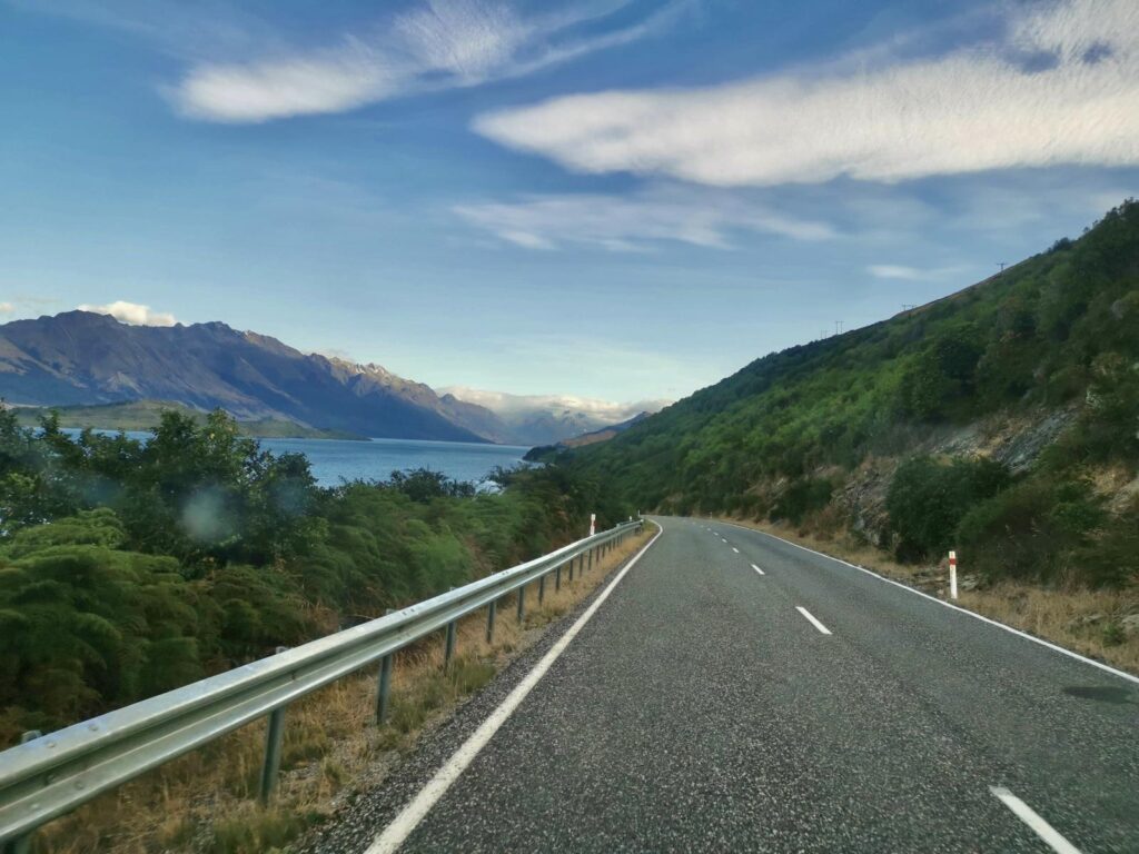 An open, winding asphalt road with a guardrail on the road to Glenorchy, showcasing Lake Wakatipu and rugged mountains under a blue sky with wispy clouds in the South Island of New Zealand.