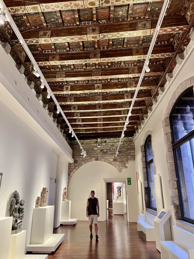 Allan standing in a bright gallery room at the Museu Etnològic i de Cultures del Món beneath an ornate medieval painted wooden ceiling.