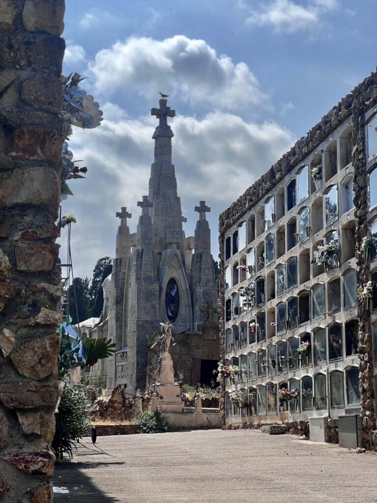 Sunlit pathway through Montjuïc Cemetery featuring terraced niches and elaborate historic stone tombs under a cloudy blue sky.