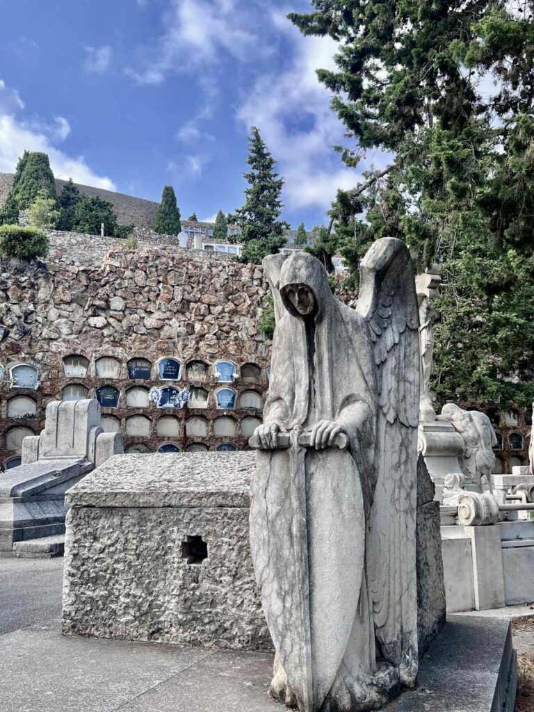 A winged angel statue resting on a stone mausoleum with hillside tombs in the background at Montjuïc Cemetery.