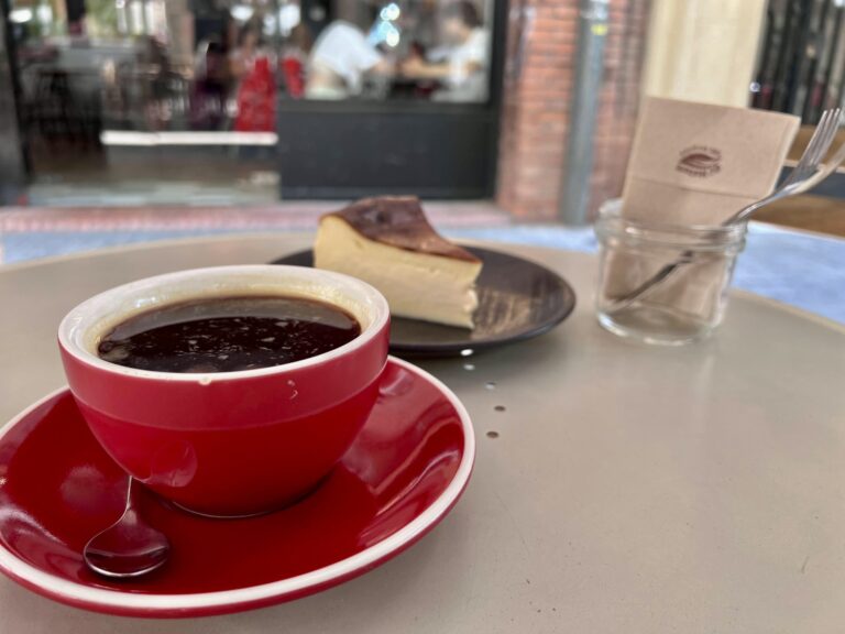 A red cup of hot coffee on a matching red saucer next to a slice of Basque burnt cheesecake on an outdoor table at Brew Coffee in Barcelona.