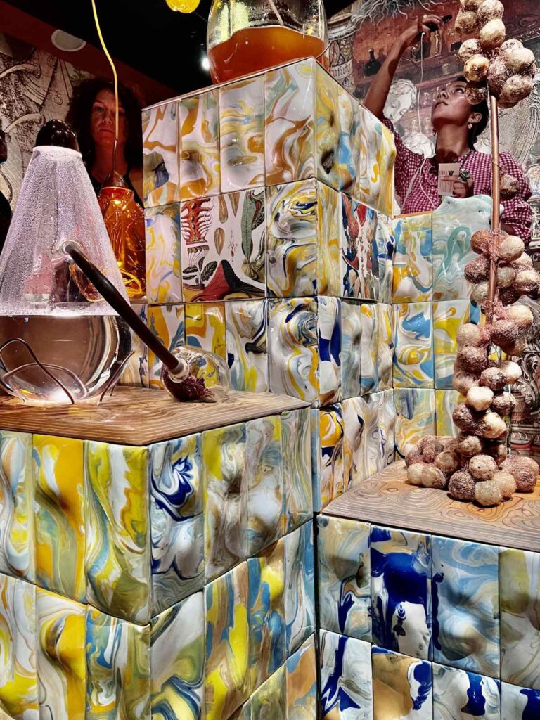 Modern alchemy and skincare artwork installation featuring colorful marbled tiles, glass retorts, and organic sculptures at the CCCB museum in Barcelona.