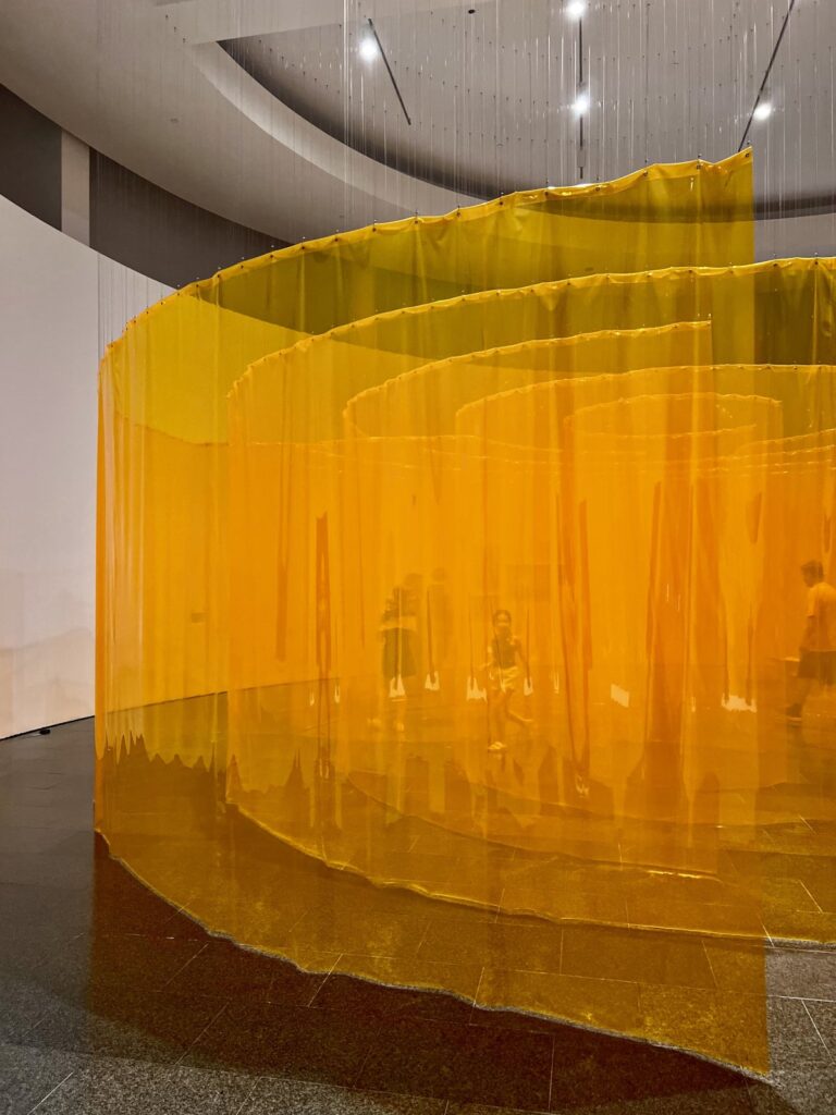 A large translucent orange mesh spiral artwork suspended from the ceiling inside a MACBA exhibition room.