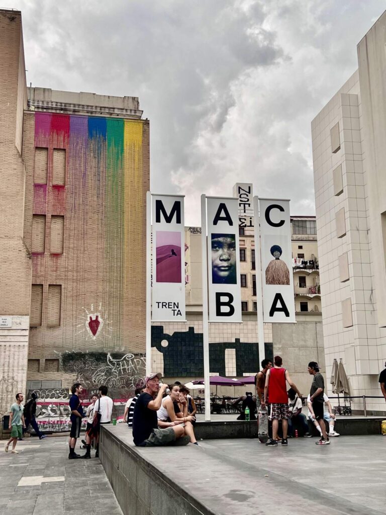 Outdoor view of the MACBA museum banners and rainbow mural with people sitting in Plaça dels Àngels plaza in Barcelona.