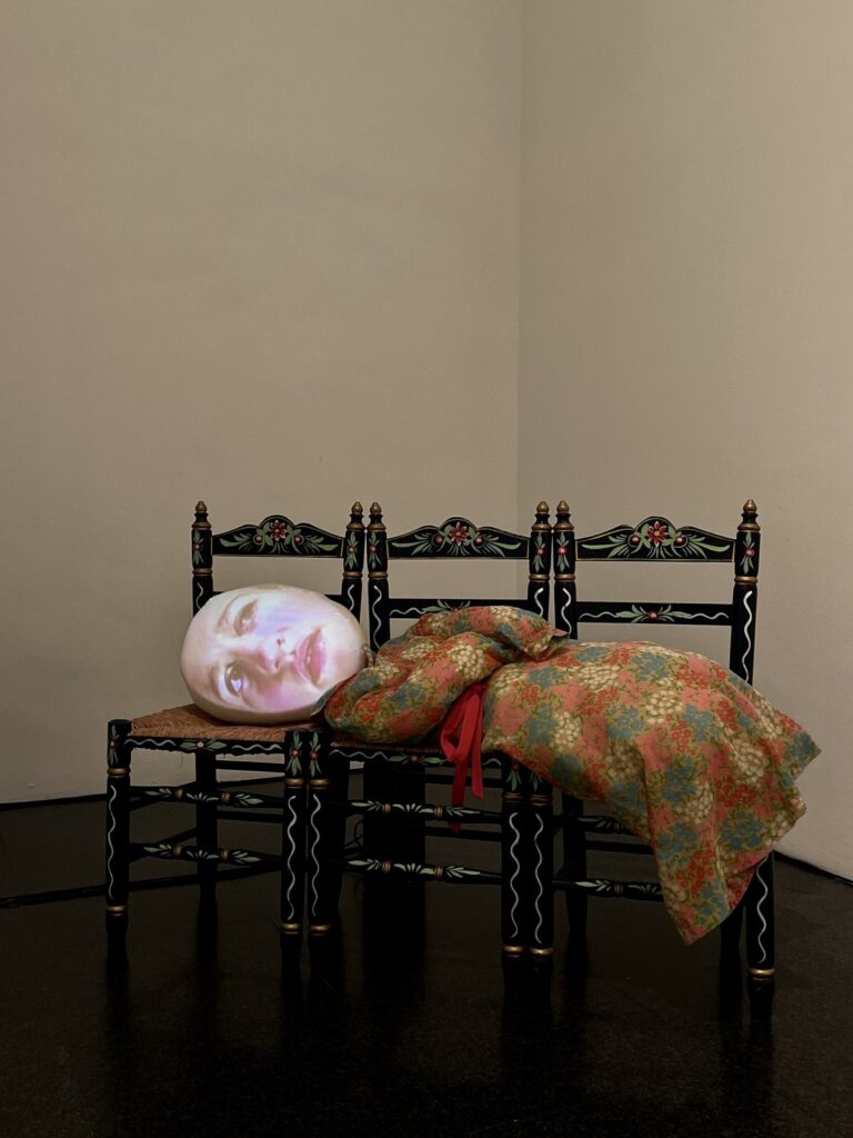 A modern art sculpture featuring a projected face on a figure lying across traditional black chairs at MACBA.