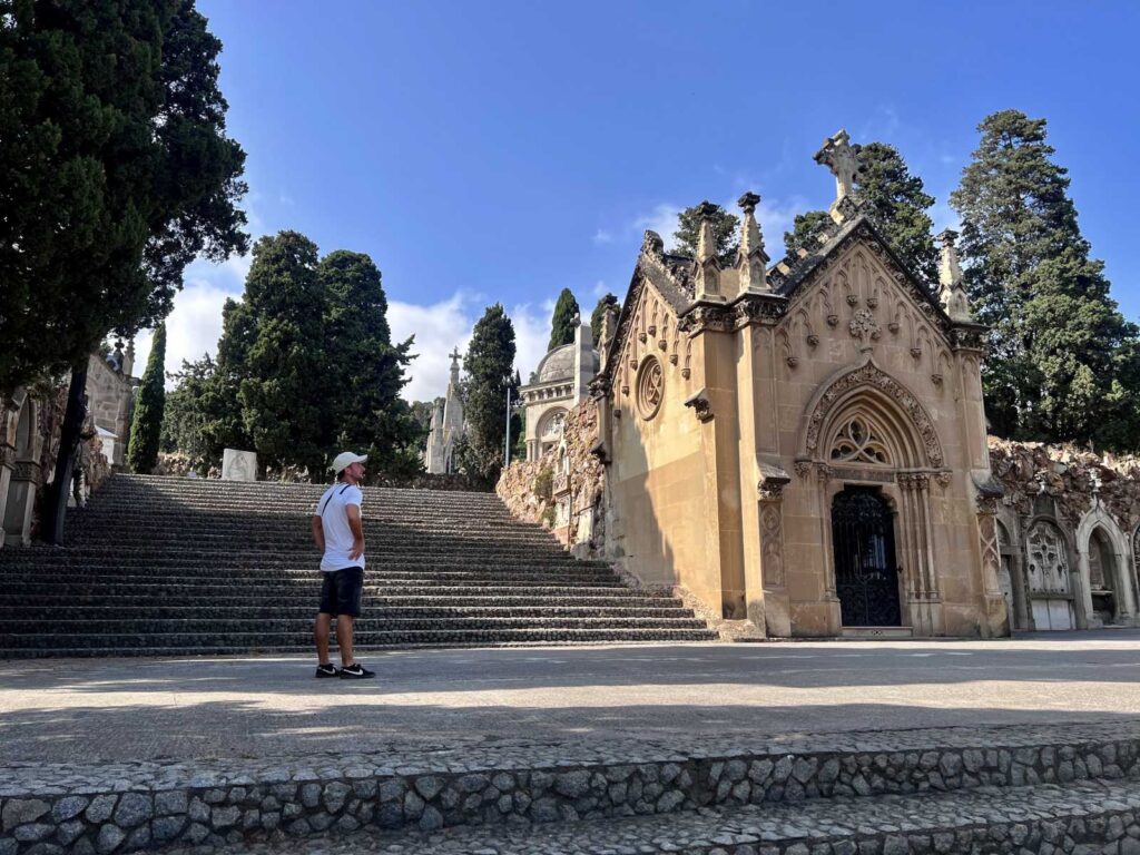 Allan looking up at a detailed stone Neo-Gothic mausoleum next to wide stairs inside Montjuïc Cemetery in Barcelona.