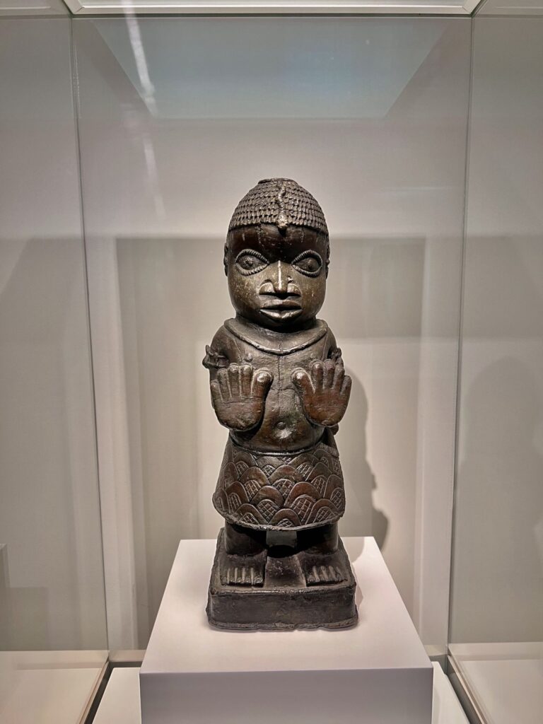 Bronze standing figure carving with raised hands on display in a glass case at the Museu Etnològic i de Cultures del Món in Barcelona.