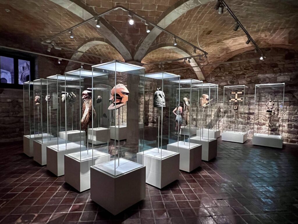 An underground stone-vaulted room in a medieval palace with glass display cases showing traditional masks at the Museu Etnològic in Barcelona.