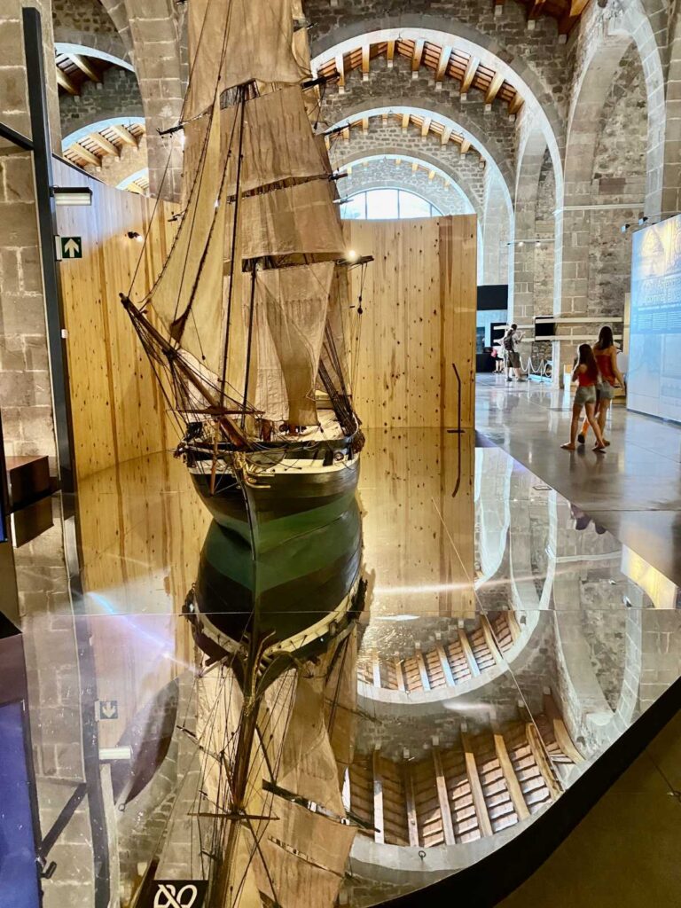 A detailed model sailing ship reflecting on a polished glass display inside the stone hall of the Barcelona Maritime Museum.