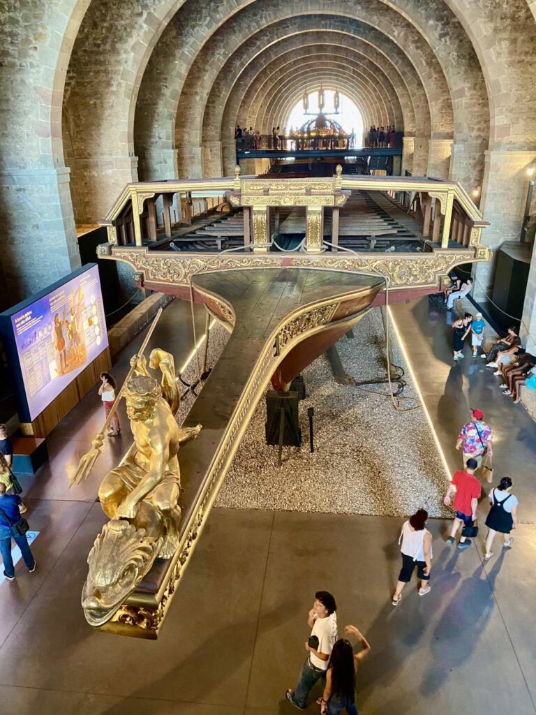 Overhead view of visitors admiring the massive ornate royal galley ship inside the stone arched hall of the Barcelona Maritime Museum.