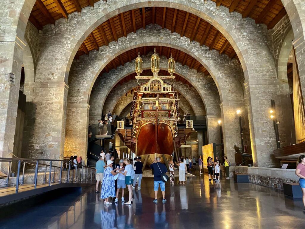 Visitors standing in front of the ornate gilded stern and golden lanterns of the Royal Galley inside the Barcelona Maritime Museum.