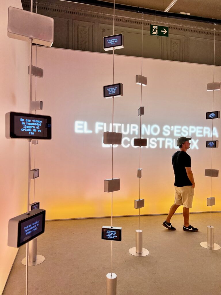 Allan walking past hanging digital display screens and glowing wall projections inside Palau Robert in Barcelona.