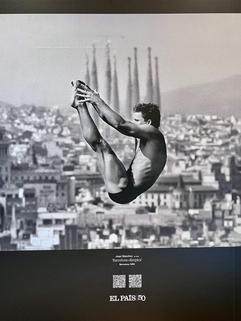A black and white photograph of an Olympic diver mid-air with the Sagrada Família in the background, displayed inside Palau Robert in Barcelona.