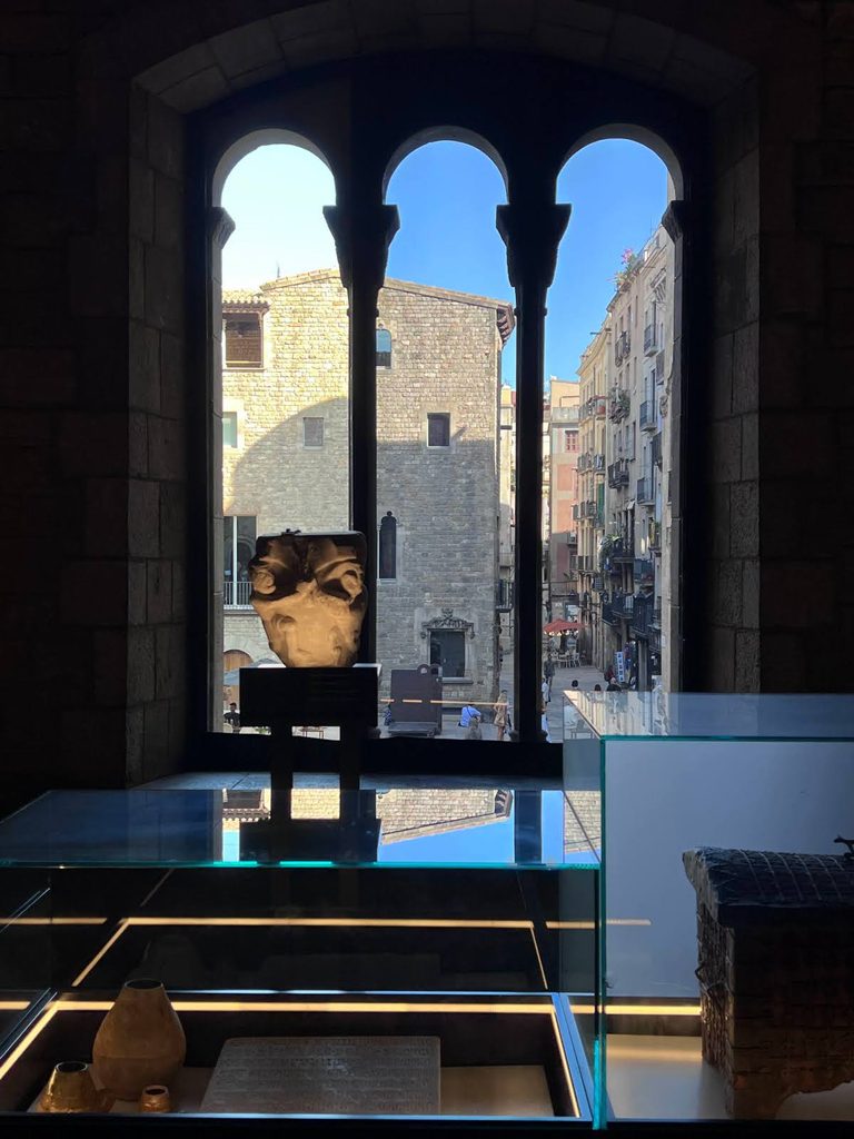 A arched gothic window inside MUHBA Plaça del Rei framing a view of historic stone buildings and a street in Barcelona's Gothic Quarter.