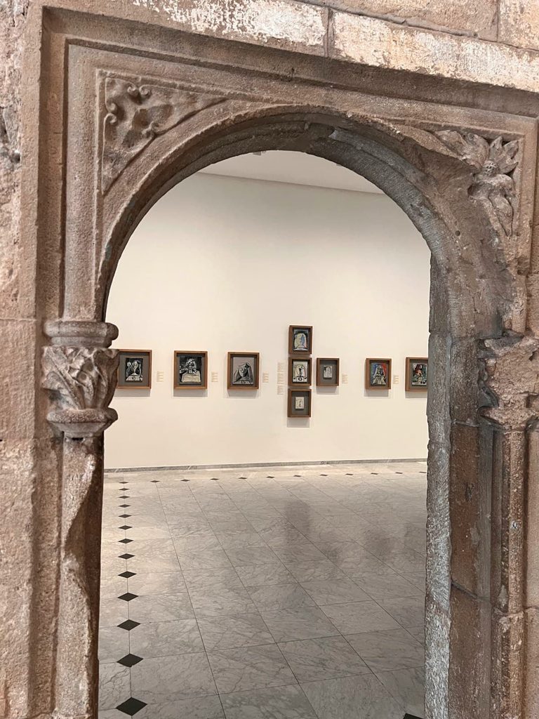 A view through a carved stone Gothic doorway into a white gallery room displaying framed artwork at the Picasso Museum in Barcelona.