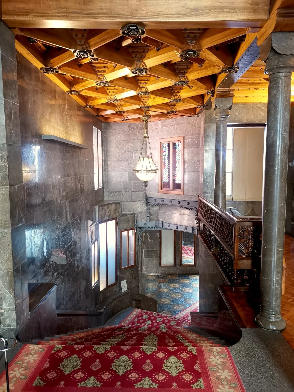 A red carpeted grand staircase surrounded by dark marble walls, columns, and an intricate wooden ceiling inside Palau Güell in Barcelona.
