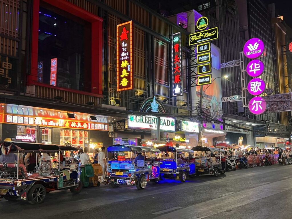 Vibrant night scene on Yaowarat Road in Bangkok Chinatown featuring glowing neon Chinese signs, street food stalls, and a line of tuk-tuks parked along the street.