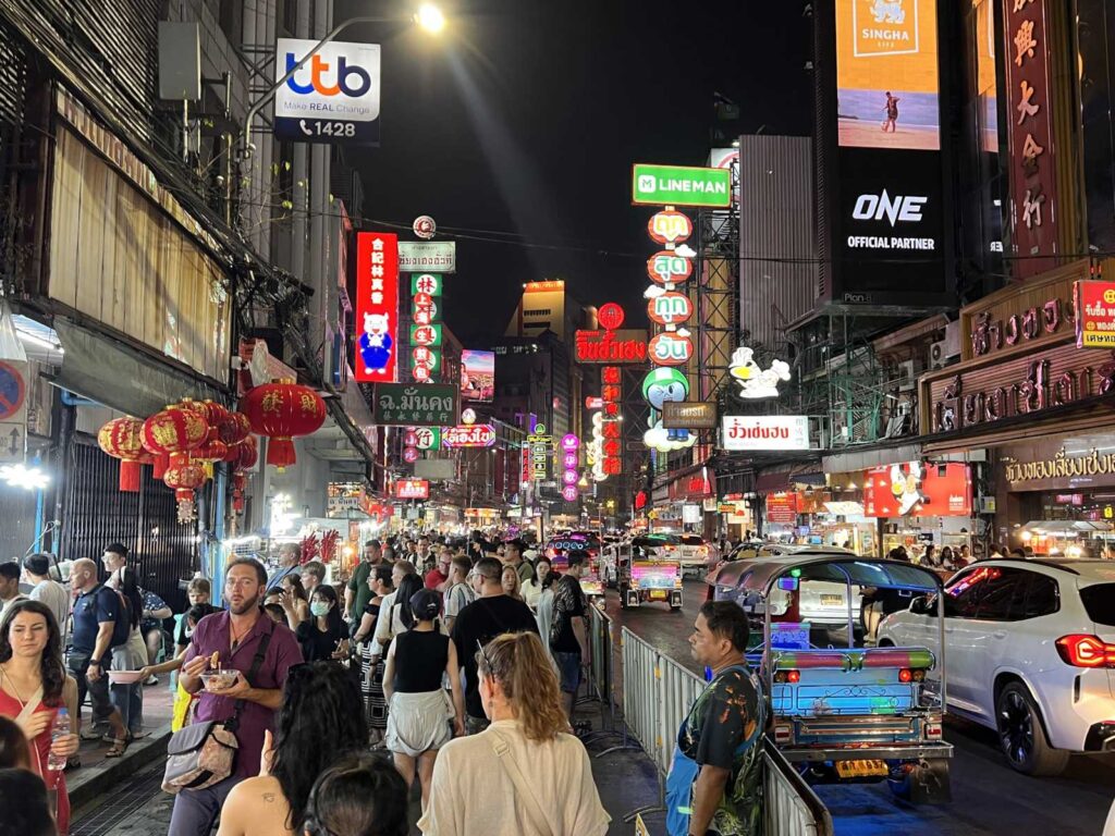 Crowded evening sidewalk packed with tourists and locals eating street food under illuminated neon signs on Yaowarat Road in Bangkok Chinatown.