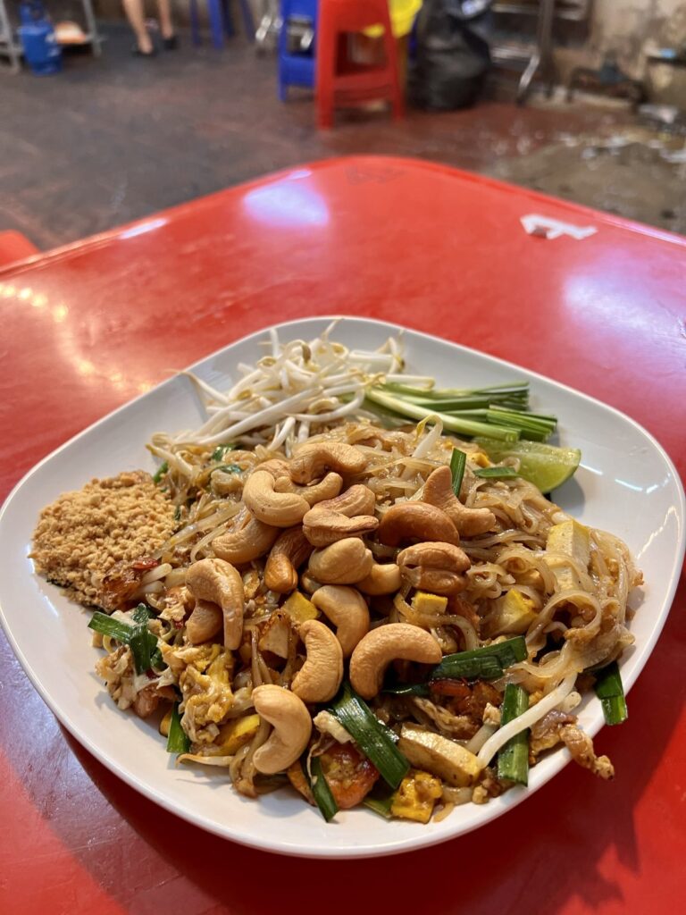 Freshly cooked Pad Thai topped with generous whole cashews, crushed peanuts, fresh bean sprouts, chives, and a lime wedge on a red table in Bangkok Chinatown.